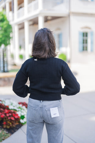 Back view showcasing the structural details of the black ribbed sweater, highlighting the soft drape of the balloon sleeves and the wide, banded hem that sits perfectly at the waistline. The sweater is paired with light grey wash denim jeans featuring classic back pockets and a visible white Vervet brand tag, emphasizing a flattering and relaxed everyday fit.