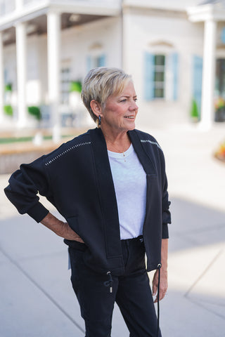 Front view of a smiling woman looking off-camera, modeling a sporty-chic black open-front jacket outdoors. The relaxed-fit layering piece features ribbed cuffs, a ribbed collar, drop shoulders accented with distinctive white dashed stitching, and a functional drawstring hem with metal toggles. She pairs the versatile jacket casually over a classic white crewneck t-shirt and black denim jeans. The everyday look is accessorized with black and gold statement hoop earrings and a delicate gold necklace.