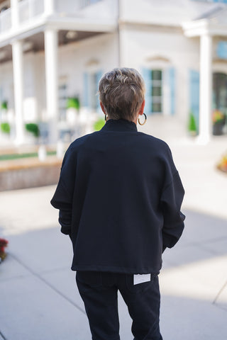 Back view highlighting the comfortable, slightly oversized drape of the black open-front jacket. The image showcases the smooth back panel, ribbed collar, and relaxed drop-shoulder silhouette that rests perfectly at the hip. The jacket is styled with black denim jeans featuring classic back pockets and a visible white sizing tag, emphasizing a casual, effortless athleisure aesthetic.