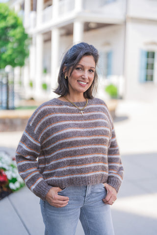 Front view of a smiling woman posing outdoors, modeling a cozy, fuzzy brown and grey striped crewneck sweater accented with thin orange and light blue horizontal stripes. The relaxed-fit knit top is styled casually with her hands resting in the pockets of light grey wash denim jeans. She accessorizes the comfortable everyday look with a gold cross pendant necklace and gold hoop earrings, set against a classic white building backdrop.