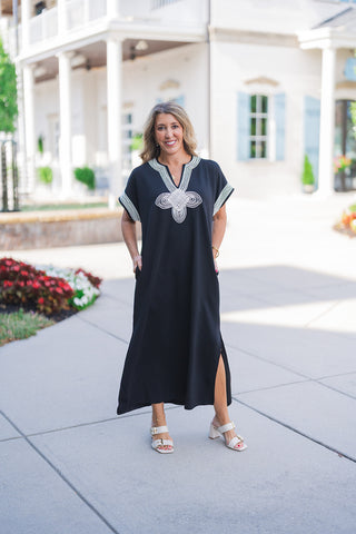 Woman smiling outdoors wearing a relaxed-fit black caftan-style midi dress featuring functional side pockets and side slits at the hem. The dress is accented with striking white geometric embroidery around a V-neckline and white striped contrast trim on the short, wide sleeves. Styled with cream double-buckle block heel sandals and delicate gold jewelry, standing on a paved walkway in front of a white building with light blue shutters.