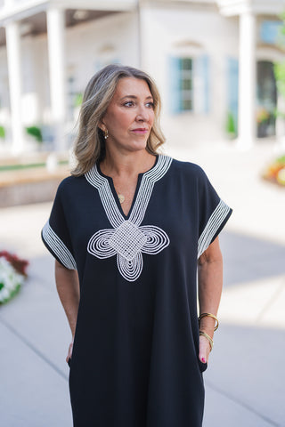 A portrait view of the black midi dress highlighting the intricate, rope-like white embroidered floral medallion and linear trim detailing that frames the V-neckline. The relaxed short sleeves feature coordinating white striped edges. The woman has her hands resting in the dress's side pockets, accessorized with layered gold pendant necklaces and small gold hoop earrings for an effortless, elevated look.