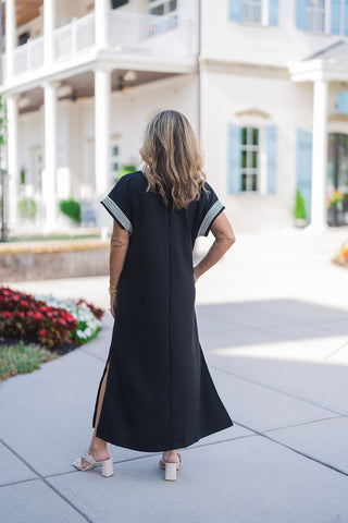 Back view of the black midi dress showcasing the flowing, comfortable silhouette, complete with a clean central back seam and knee-high side slits that allow for easy movement. The white striped trim on the wide sleeves is clearly visible from behind. The look is finished with cream block-heel sandals, emphasizing the dress's versatile, warm-weather appeal.
