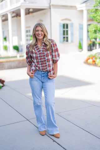 Full body front view of a smiling blonde woman posing outdoors, modeling a transitional fall outfit featuring a short-sleeve plaid top in shades of red, navy blue, and cream. The blouse features voluminous puff sleeves, a V-neckline, and a feminine ruffle detail cascading down the front button placket. She pairs the top with light-wash blue denim jeans featuring retro-inspired utility front patch pockets and a subtle flared leg. The look is completed with tan suede pointed-toe boots, a chunky gold chain nec