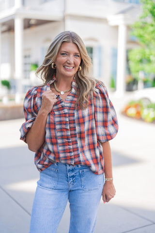 A portrait view highlighting the textured details and flattering fit of the plaid puff-sleeve top. The close-up showcases the rich red, navy, and cream plaid pattern, the structured puff sleeves with gathered shoulders, and the delicate ruffle trim framing the center button placket. The V-neckline perfectly frames a trendy chunky gold chain necklace, complemented by thick gold hoop earrings for an elevated everyday style.