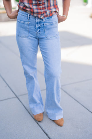 Lower body front view of a woman modeling light wash blue denim jeans featuring retro-inspired utility front patch pockets. The flattering wide-leg silhouette is finished with a prominent, thick bottom hem. The high-rise jeans are styled casually with a tucked-in orange, red, and blue plaid button-down shirt and brown suede pointed-toe shoes, photographed outdoors on a paved walkway.