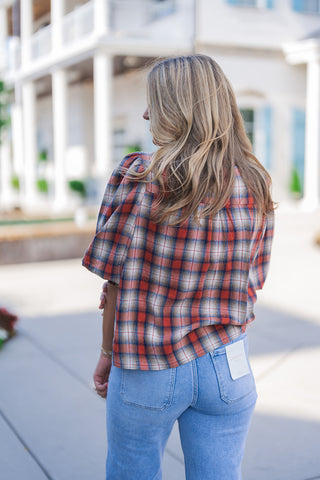 Back view of the plaid puff-sleeve top showcasing its comfortable, relaxed drape and soft pleating along the back yoke. The image highlights the continuous woven plaid pattern across the back and sleeves. The blouse rests effortlessly over light-wash blue denim jeans, which feature classic back patch pockets and a visible white Lovervet brand tag attached to the right pocket, emphasizing a stylish, trendy aesthetic.