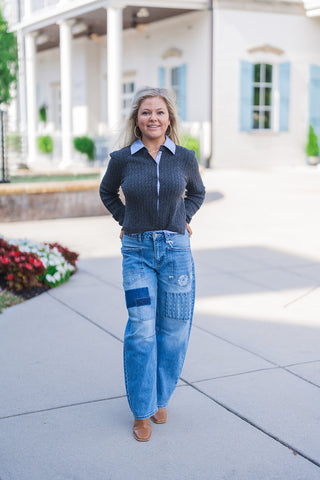 Full body front view of a smiling blonde woman posing outdoors, modeling a preppy, layered fall outfit. She wears a dark grey ribbed knit cardigan buttoned casually over a crisp light blue collared shirt. The layered tops are paired with trendy, wide-leg medium-wash blue denim jeans featuring distinct patchwork squares, subtle distressing, and utility details. She finishes the stylish everyday look with tan leather square-toe boots and large gold hoop earrings, standing on a paved walkway in front of a whit