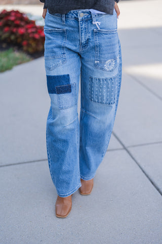 Lower body front view highlighting the unique novelty details of medium-wash blue denim jeans. The high-waisted, wide-leg pants feature retro-inspired utility front patch pockets and intricate patchwork designs on the thighs, including a contrasting dark wash denim patch, a distressed stitched rectangular patch, and subtle floral embroidery near the left pocket. Distinct horizontal seams cross just below the knees, creating a trendy barrel-leg silhouette. The denim drapes comfortably over tan leather square