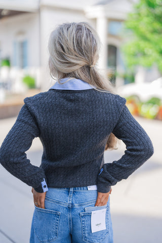 Back view of the layered outfit showcasing the tailored, comfortable fit of the dark grey ribbed cardigan. The image highlights the sweater's subtle shoulder structure and the light blue shirt collar and sleeve cuffs peeking out for a polished, preppy contrast. The cardigan rests cleanly at the waistband of the medium-wash blue jeans, which feature classic back patch pockets and visible white Kate and Vervet brand sizing tags, emphasizing a trendy, texture-rich aesthetic.