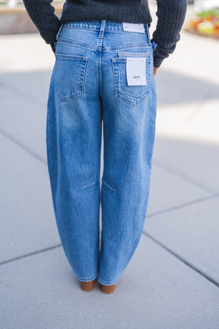 Lower body back view of the medium-wash patchwork jeans, showcasing their comfortable, relaxed fit and distinct curved barrel-leg shape that tapers slightly at the ankle. The jeans feature classic back patch pockets, standard belt loops, and unique horizontal seaming behind the knees. A white Vervet brand tag is visibly attached to the right back pocket, alongside a Kate style tag on the waistband. The jeans pair effortlessly with a dark grey ribbed knit top and tan leather boots for a chic, textured everyd