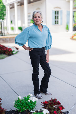 Woman posing outdoors in the Cornflower Collar Blouse, a light cornflower blue woven top with a ruffled mandarin collar, a split neck V-placket, and dramatic 3/4-length balloon sleeves with ruffled cuffs. Styled for an elevated casual look with black slim-fit jeans, western boots, and black-and-gold hoop earrings in front of a white building with light blue shutters.