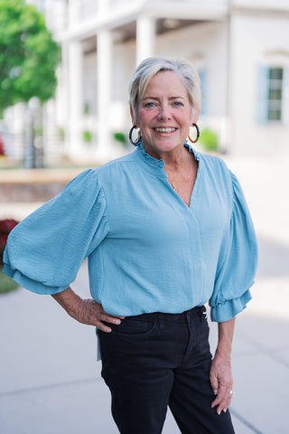 A portrait view of the Cornflower Collar Blouse highlighting the delicate ruffle detail trim along the collar and open split neckline. Demonstrates the soft texture of the cornflower blue fabric, dropped shoulder seams, and voluminous puff sleeves tucked front-wise into black denim.