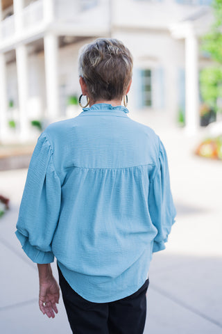 Back view of the Cornflower Collar Blouse showcasing the subtle ruffle along the back collar and a horizontal back yoke seam with light gathering for a fluid, relaxed fit. Highlights the extended curved high-low hem draped comfortably over black jeans.