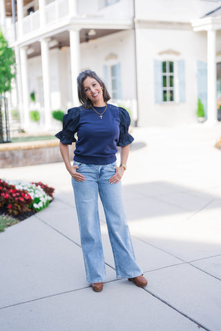 Full body front view of a smiling woman posing outdoors with her hands in her pockets, modeling a unique navy blue mixed-media top. The elevated casual shirt features a comfortable soft knit bodice contrasted with structured dark wash denim statement puff sleeves. She pairs the trendy top with light-wash blue wide-leg denim jeans featuring subtle rhinestone side embellishments. The outfit is completed with brown leather clogs, a gold cross pendant necklace, and a gold watch, set against a classic white buil