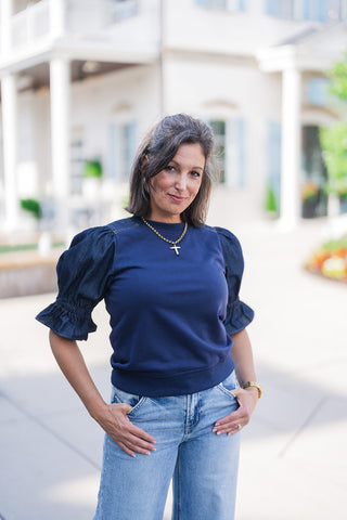 A portrait view highlighting the architectural and textural details of the navy denim puff sleeve top. The close-up showcases the striking mixed-media design, emphasizing the contrast between the smooth navy blue knit bodice and the voluminous dark denim sleeves, which feature gathered shoulders and elastic ruffled cuffs. A classic ribbed crew neckline frames a delicate gold cross necklace, offering a chic, elevated take on an everyday basic.