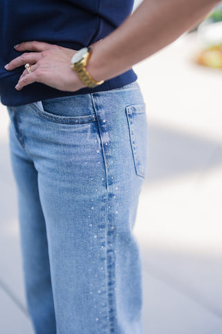 A detailed close-up side view highlighting the hip and pocket construction of light wash blue denim jeans. The design features delicate, sparkling rhinestone crystal embellishments cascading down the outer side seam, adding a glamorous touch to the classic denim. The model has her hand resting on her hip, wearing a navy blue top and a gold watch, showcasing a stylish and elevated casual look.