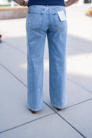 Lower body back view modeling the light wash blue denim jeans, showcasing their comfortable, wide-leg silhouette. The jeans feature classic back patch pockets, standard belt loops, and a visible white Flying Monkey brand tag attached to the right back pocket. The relaxed-fit denim drapes cleanly over brown shoes, paired effortlessly with a navy blue top for a trendy, everyday aesthetic.