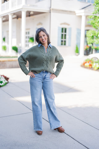 Full body front view of a woman smiling with her hands on her hips, modeling a chunky olive green V-neck sweater featuring a prominent central cable-knit design and ribbed texture. The sweater is styled in a trendy layered look over a classic blue denim collared shirt. She completes the casual, cozy aesthetic with light-wash, wide-leg jeans featuring subtle side-seam stud detailing and brown leather platform clogs, standing outdoors on a paved walkway.