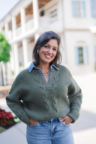 A portrait view of the woman highlighting the rich textures of the olive green V-neck sweater layered over a crisp blue denim shirt. The close-up showcases the sweater's central cable-knit braiding and ribbed raglan sleeves. Her hands rest casually in the front pockets of her light-wash jeans, and the neckline is accessorized with a gold cross pendant necklace and classic gold hoop earrings for an elevated everyday look.