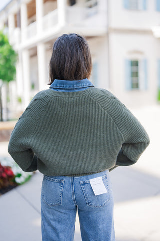 Back view showcasing the relaxed, slightly cropped drape of the olive green ribbed knit sweater and its raglan sleeve construction, with the blue denim collar smartly popping out at the neckline. The sweater rests comfortably at the waistline of the light blue wash denim jeans, which feature classic back pockets and a visible Flying Monkey Los Angeles brand tag.