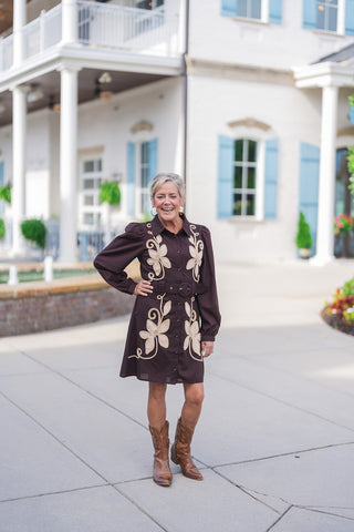 Woman posing outdoors in the Vintage Prairie Espresso Dress, a long-sleeve, button-front mini dress in a rich espresso brown hue featuring prominent beige felt floral applique and cord embroidery along the bodice and skirt. Designed with slight puff shoulders, a spread collar, and a matching fabric grommet belt that cinches the waist. Styled for a western-chic look with brown leather cowboy boots and gold hoop earrings in front of a white building with light blue shutters.