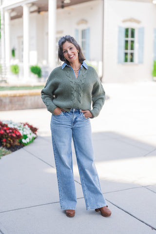 Full body front view of a smiling woman modeling a cozy layered fall outfit outdoors. She is wearing a chunky olive green V-neck sweater featuring a prominent central cable-knit pattern and ribbed texture, smartly layered over a classic blue denim shirt. Her hands are resting casually in the front pockets of light-wash, wide-leg denim jeans that feature subtle side-seam stud embellishments. The everyday look is completed with brown leather platform clogs and a gold cross necklace, set against a classic whit
