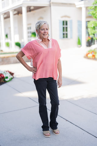 Woman posing outdoors in the Sweet Peach Puff-Sleeve Top, a vibrant peach-pink relaxed pullover blouse featuring a split V-neckline framed with cream crochet lace trim and a contrast striped collar. The top features wide cap sleeves with folded cuffs and subtle cream contrast lace side panels. Styled for an everyday casual look with black straight-leg denim jeans, platform wedge sandals, and dark hoop earrings in front of a white building with light blue shutters.