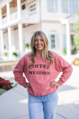 Full body front view of a smiling blonde woman posing outdoors with her hands on her hips, modeling a cozy dusty pink crewneck sweatshirt. The front of the pullover features the phrase "COFFEE WEATHER" in brown, textured applique lettering. The relaxed-fit top is styled casually with light wash blue denim jeans featuring retro-inspired utility front patch pockets. She accessorizes the comfortable everyday look with gold hoop earrings and a gold chain necklace, set against a classic white building with light