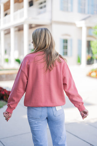Back view of the dusty pink crewneck sweatshirt highlighting its comfortable, relaxed drop-shoulder drape, ribbed cuffs, and ribbed bottom hem. The cozy fall pullover rests easily over a pair of light wash blue denim jeans, which showcase classic back patch pockets and a visible white brand tag on the right side, emphasizing an effortless and trendy seasonal aesthetic.