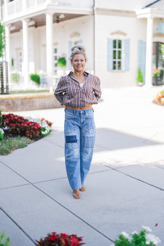 Woman smiling playfully while modeling a casually chic outfit, featuring a brown, blue, and white vertical striped button-down shirt tucked up to reveal a sliver of midriff over a white tank top. She is wearing trendy medium-wash wide-leg denim jeans adorned with textured denim patchwork squares and front utility pockets. The look is completed with brown square-toe boots, gold hoop earrings, and a relaxed high bun, set against an elegant white building with columns and blue shutters.