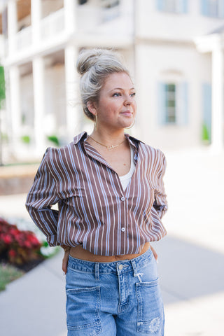 A waist-up view highlighting the relaxed, effortless styling of the brown and blue striped long-sleeve button-down shirt, worn unbuttoned at the top over a classic white undershirt. This closer crop showcases the front utility pockets of the denim jeans and the model's accessories, which include large gold hoop earrings and a delicate gold script nameplate necklace.