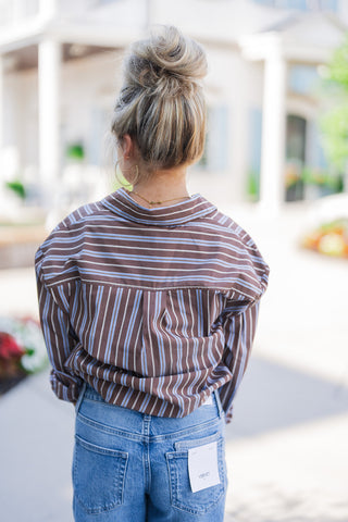 Back view of the outfit demonstrating the relaxed drape and horizontal back yoke pattern of the striped button-down shirt. The image captures the comfortable, loose fit of the top paired with the structured waist of the medium-wash jeans, which feature classic back pockets with a visible white brand tag on the right side. The model is facing away, highlighting her voluminous blonde high bun hairstyle.
