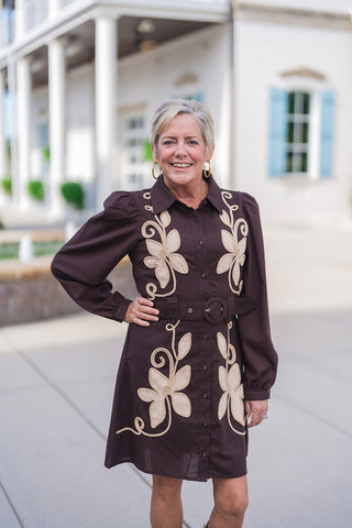 A portrait view of the Vintage Prairie Espresso Dress highlighting the bold beige cream floral applique motifs bordered with matching soutache cord trim. Demonstrates the structured spread collar, button-down front closure, subtle gathered shoulder seam details, and the covered circular buckle on the wide waist belt.