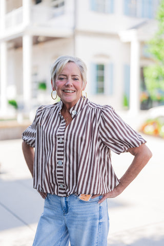 Woman smiling and posing with hands on her hips, modeling a brown and white vertically striped short-sleeve blouse. The top features a contrasting horizontal striped button placket, large round statement buttons, a band collar, and softly gathered puff sleeves. The relaxed, elevated top is styled with casual light-wash denim jeans, a delicate gold necklace, and large gold hoop earrings for a breezy, everyday aesthetic outside a classic white building.