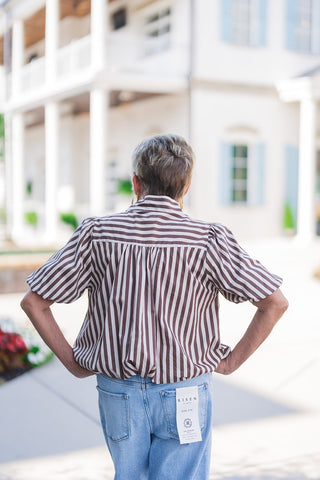 Back view of the brown and white striped blouse highlighting the structural detail of a horizontal striped back yoke with soft gathering beneath it. The tailored yet airy design creates a comfortable, blouson drape over light-wash blue jeans. The denim features classic back pockets with a visible Risen Los Angeles brand tag, showcasing a relaxed but flattering fit.