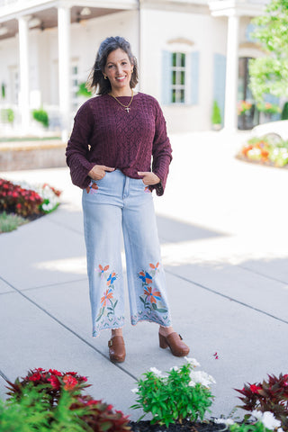 Full body front view of a smiling woman posing outdoors, modeling a cozy burgundy pointelle-knit sweater with a subtle front tuck. She pairs the long-sleeve top with light-wash, wide-leg cropped jeans that feature vibrant, multi-colored floral embroidery climbing up the lower legs and a smaller floral accent near the right front pocket. The outfit is styled for a casual, bohemian-chic aesthetic with a gold cross necklace and brown leather heeled buckle clogs, set against a classic white building and a flowe