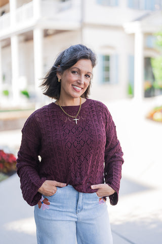 A portrait view of the Mulled Wine Pointelle Sweater highlighting the delicate pointelle knit texture, ribbed crew neck, and rich deep wine shade. The relaxed fit sweater is tucked subtly into embroidered light-wash denim, accessorized with a layered gold chain and small gold hoop earrings.