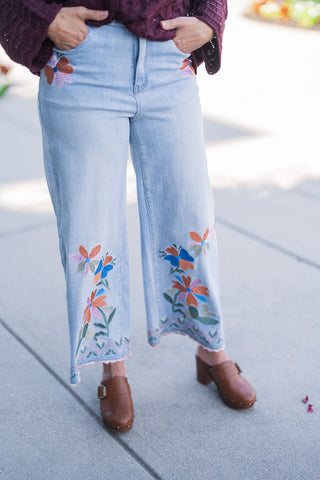Lower body front view focusing on the fit and intricate details of the light-wash denim. The image highlights the high-waisted, wide-leg crop silhouette and the striking floral embroidery featuring orange, blue, and purple blooms with green foliage. The unique geometric zigzag stitching and delicate peach-toned scalloped edge of the hemline are clearly visible, draped perfectly over brown leather platform buckle clogs.
