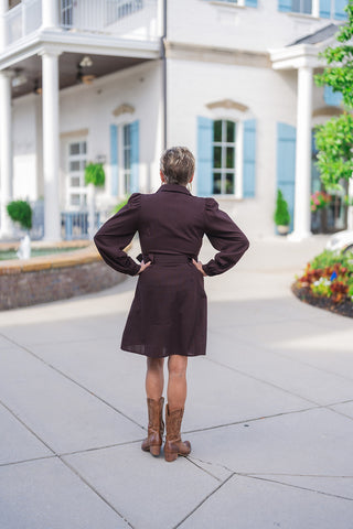 Back view of the Vintage Prairie Espresso Dress showing the clean, unembellished dark brown back panel and A-line mini skirt fit. Highlights the full long sleeves with subtle puff shoulders, buttoned cuffs, and the back profile of the matching waist belt with the model's hands placed on her hips.
