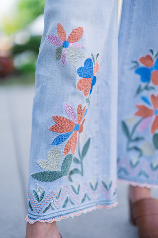 A macro close-up view highlighting the high-quality craftsmanship of the denim's embroidery. The detailed shot showcases the rich texture of the heavy threadwork used to create the vivid orange, blue, and purple flower petals and interlocking green leaves. It also provides a clear view of the hem's intricate finishing, featuring green and orange geometric zigzag stitches bordered by a precision-cut, scalloped edge.