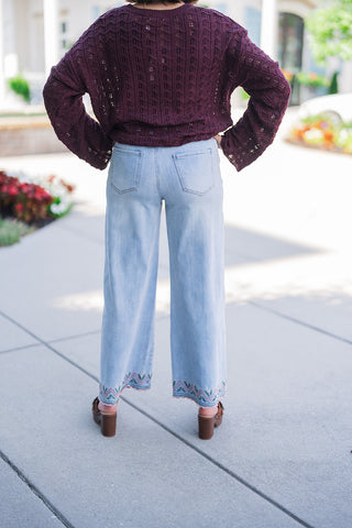 Back view from the waist down, showcasing the clean, flattering rear fit of the light-wash wide-leg jeans with classic back patch pockets. The image highlights how the geometric, color-stitched scalloped hem continues around the back of the pant legs. The burgundy open-knit sweater rests comfortably at the waistline, completing the effortless, texture-rich transitional fall outfit.