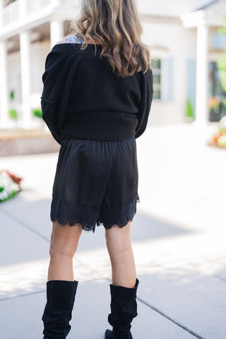 Back view of the black satin lace-trim shorts showcasing the relaxed, fluid drape of the silky fabric and the intricate detailing of the scalloped eyelash lace along the bottom hem. The elevated, evening-ready shorts are styled with a black ribbed-hem sweater draped effortlessly off the shoulders and black suede slouchy boots for a chic, texture-rich aesthetic set against an outdoor paved walkway.