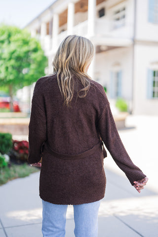 Back view highlighting the relaxed, slightly oversized fit of the brown textured knit cardigan. The image showcases the soft, boucle-like fabric and the self-tie belt resting securely across the back waist. The cardigan falls elegantly below the hips, paired casually with light-wash blue denim jeans for an effortless, everyday outfit.