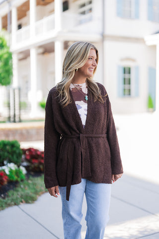 Front view of a smiling blonde woman looking off-camera, modeling a cozy brown textured knit cardigan featuring a classic tie-waist belt. The open-front sweater is elegantly layered over a brown and white abstract print top and styled with light-wash blue jeans. She accessorizes the comfortable fall look with gold hoop earrings and a layered gold necklace featuring a turquoise pendant, standing outdoors on a paved walkway.