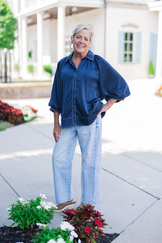 Woman posing outdoors in the Ocean Blue Satin Blouse, a relaxed deep blue satin button-down top featuring contrast light-colored zig-zag accent stitching along the placket, collar, hem, and back seam. The top boasts voluminous three-quarter length balloon sleeves and a spread collar, styled effortlessly with light-wash wide-leg denim jeans, platform sandals, and gold hoop earrings.