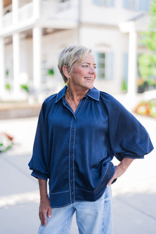 A portrait view of the Ocean Blue Satin Blouse highlighting the subtle sheen of the navy satin fabric and the detailed light blue contrast pick-stitching framing the placket, lapels, and hem. The top features a relaxed v-neck opening, covered buttons, and draped raglan-style balloon sleeves with fitted cuffs.