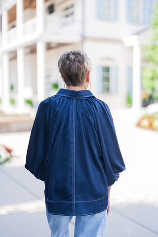 Back view of the Ocean Blue Satin Blouse showcasing the dramatic vertical center seam defined by contrast light blue stitching and delicate gathers radiating from the back yoke. Demonstrates the loose, airy drape of the tunic length and wide billowy sleeves over light-wash jeans.