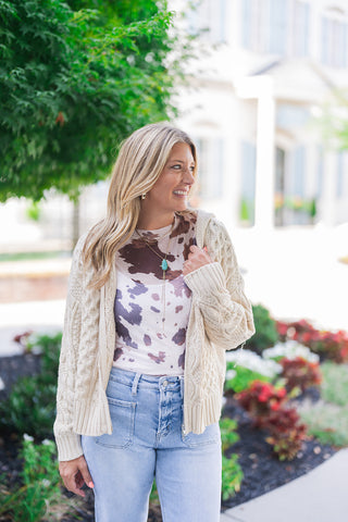 Front view of a smiling blonde woman looking off-camera, modeling a cozy cream cable-knit hooded cardigan worn open. The highly textured sweater is layered over a trendy brown and white cow print top and paired with light-wash blue denim jeans featuring utility front patch pockets. She accessorizes the casual, transitional outfit with gold hoop earrings and a layered gold necklace with a turquoise pendant, standing outdoors against a soft landscaped background.