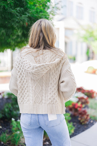 Back view of the cream hooded cardigan showcasing its rich texture, featuring intricate diamond and classic cable knit patterns down the back and sleeves. The image highlights the sweater's relaxed, slightly cropped fit, cozy attached hood, and thick ribbed bottom hem. It is styled with light-wash blue denim jeans displaying classic back patch pockets and a visible white brand tag, emphasizing a comfortable everyday aesthetic.